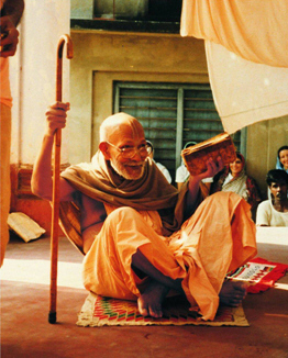 Srila Bhakti Promode Puri Goswami Maharaja seated outdoors holding his sannyasa danda, with devotees nearby