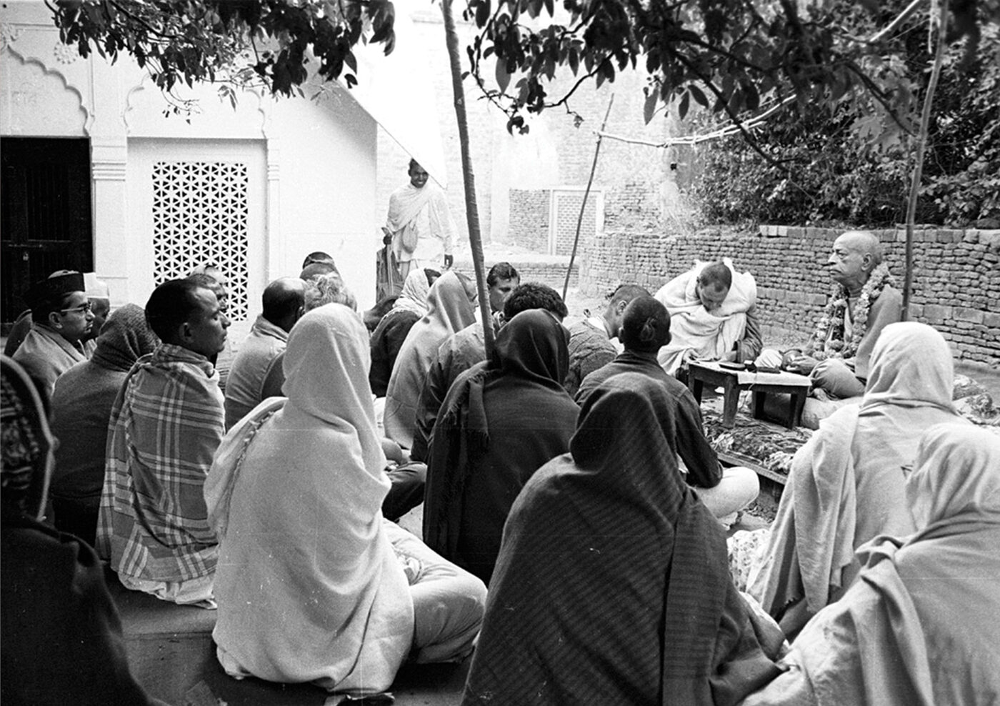 Srila Prabhupada giving an outdoor lecture to a seated audience in Vrindavan, near the samadhi of Rupa Gosvami.