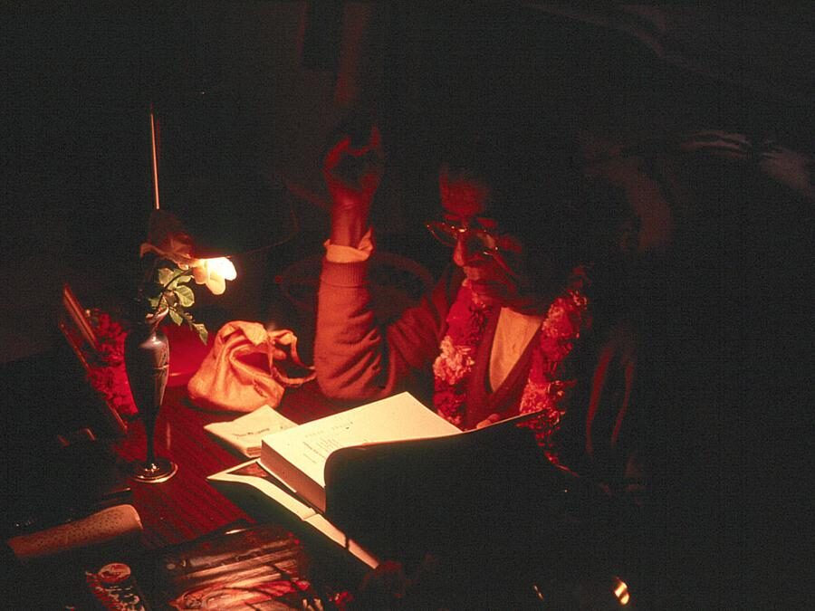Srila Prabhupada absorbed in study and writing by lamplight, a book open before him.