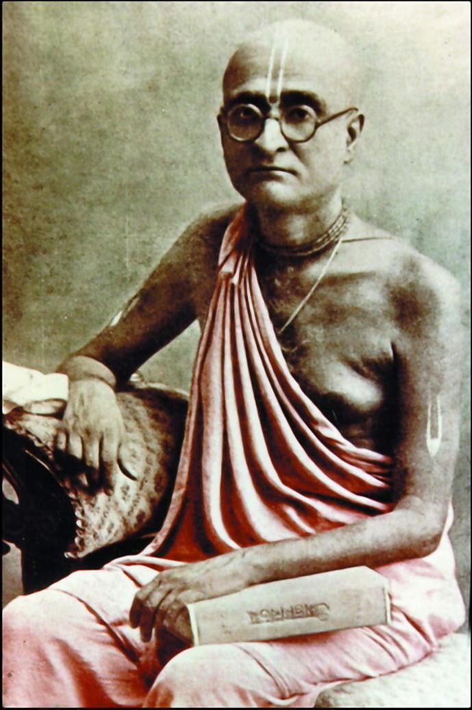 Bhaktisiddhanta Sarasvati Thakura seated, wearing round glasses, resting hand on a stack of books -- a well-known, widely reproduced portrait
